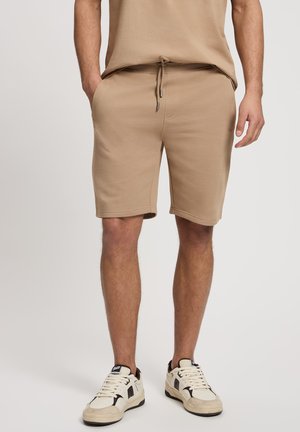 Beżowe bawełniane szorty z elastycznym pasem, sznurkiem, bocznymi kieszeniami i luźnym krojem, w zestawie z białymi i brązowymi trampkami.