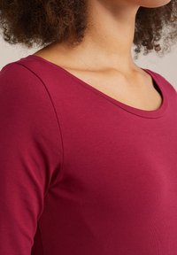 Burgunderfarbenes Langarm-T-Shirt mit Rundhalsausschnitt, aus weichem Baumwollstoff und gerippter Textur. Nahtdetails entlang der Schultern.