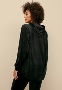 Zwarte hoodie in een zachte, fluwelen stof, met een relaxte pasvorm, lange mouwen en een afgeronde zoom. Subtiele glans textuur.