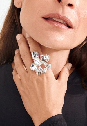 Vrouw met een lichte huid die een grote zilveren ring in bloemvorm aan haar linkerhand draagt, haar nek aanraakt, zwarte top met lange mouwen zichtbaar.