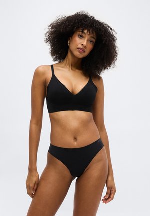 Γυναίκα με σγουρά μαλλιά που φοράει μαύρο στηθόδεσμο και εσώρουχα, στέκεται μπροστά σε μια απλή ανοιχτή φωτεινή επιφάνεια με ουδέτερη έκφραση.