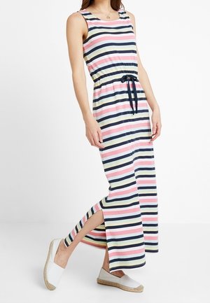 Femme portant une robe sans manches jusqu'aux chevilles avec des rayures horizontales roses, bleu marine, blanches et jaunes, une fente latérale, une ceinture bleu marine et des espadrilles blanches.