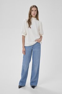 Ženska, oblečena v belo majico z nabranimi rokavi in široke modre jeans hlače, stoji z eno roko v žepu na enobarvnem belem ozadju.