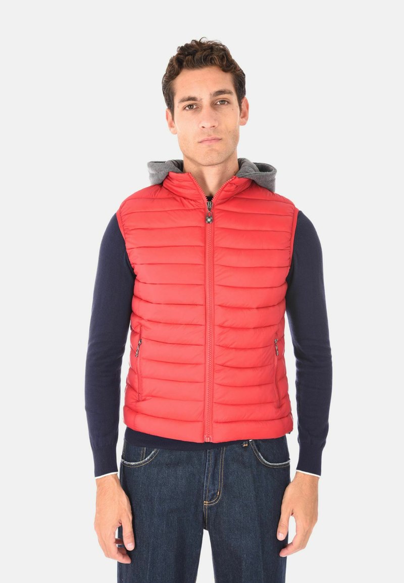 Uomo che indossa un gilet imbottito rosso senza maniche con zip frontale sopra una camicia a maniche lunghe blu navy e jeans blu, in piedi di fronte a uno sfondo bianco.