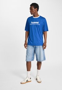 Kungligt blå kortärmad tröja med vertikala ränder och vit "hummel PERFORMANCE"-logotyp, kombinerad med ljusa denimshorts och sneakers.