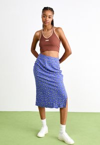 Brun halterneck crop topp og blå midi-skjørt med hvit blomsterdesign. Skjørtet har en sidespleis. Modellen har på seg hvite joggesko og sokker.