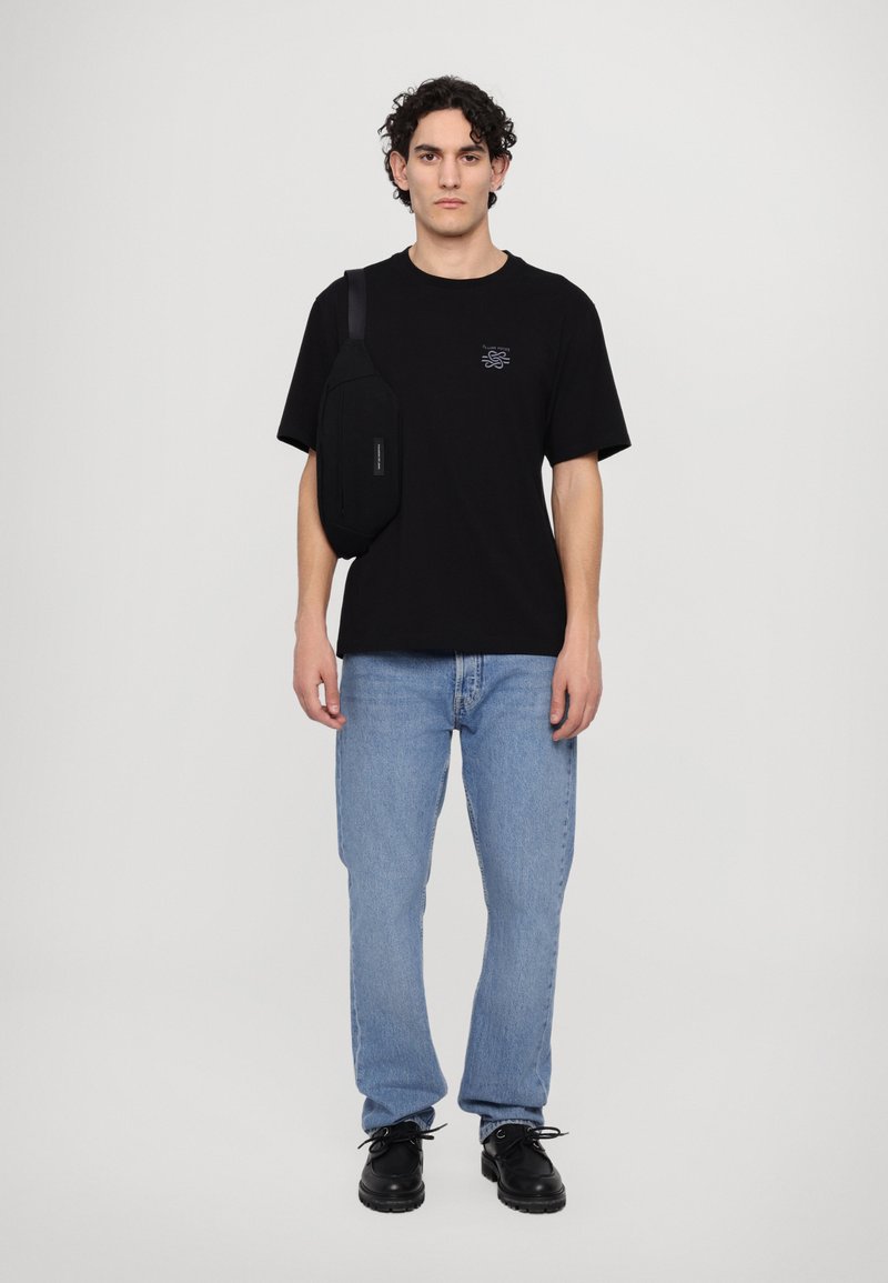 Black short-sleeve t-shirt with small graphic, blue denim jeans, black slip-on shoes, and a black shoulder bag worn across the body.