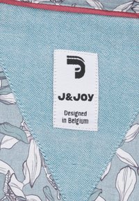 Witte kledinglabel met zwart "J&JOY" logo en de tekst "Ontworpen in België" op een blauwe bloemenstof achtergrond.