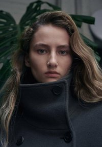 Cappotto in lana grigio carbone con collo alto, dotato di bottoni neri e design pulito. Il modello ha capelli lunghi e mossi e occhi verdi.