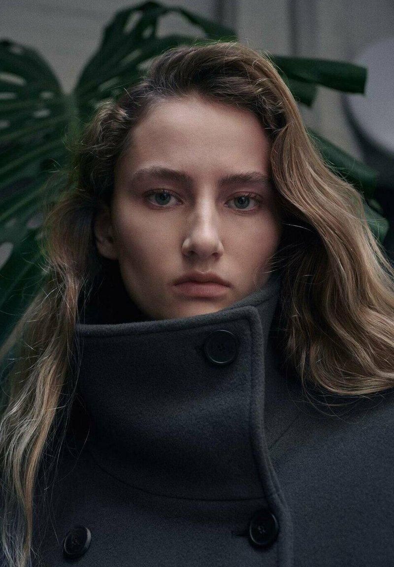 Cappotto in lana grigio carbone con collo alto, dotato di bottoni neri e design pulito. Il modello ha capelli lunghi e mossi e occhi verdi.