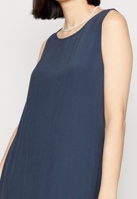 Nainen, joka on pukeutunut hihattomaan tummansiniseen mekkoon ja yksiriviseen helmikaulaketjuun, lyhyet tummat hiukset ja vaalea iho.