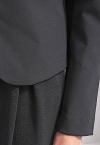 Veste noire avec un col arrondi en gros plan, associée à une jupe en tissu noir texturé. Matériau lisse et coupe ajustée.