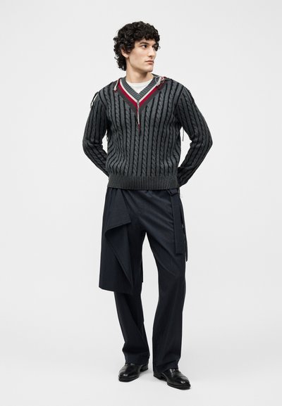 Homme aux cheveux bouclés portant un pull en tricot câblé gris foncé avec bordure rouge et un pantalon noir à rayures fines avec un détail de tissu superposé, chaussures noires.