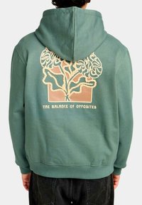 Sudadera verde con un gran gráfico en la parte posterior que presenta un diseño de árbol en crema y marrón, con el texto "EL EQUILIBRIO DE LOS OPUESTOS" debajo.