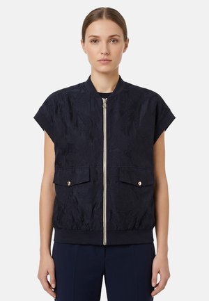 Donna che indossa una giacca blu navy a maniche corte con chiusura lampo e due tasche frontali con patta, in piedi davanti a uno sfondo bianco uniforme.