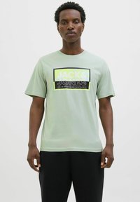 Lysegrøn bomulds T-shirt med et dristigt sort og neongrafisk logo "JACK & JONES" i rektangulært design. Korte ærmer, rund hals.