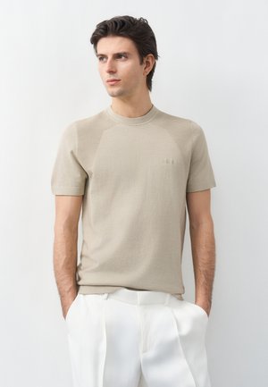 Ung mand iført en beige kortærmet striktrøje og hvide bukser, stående med hænderne i lommerne og kigger let mod venstre.