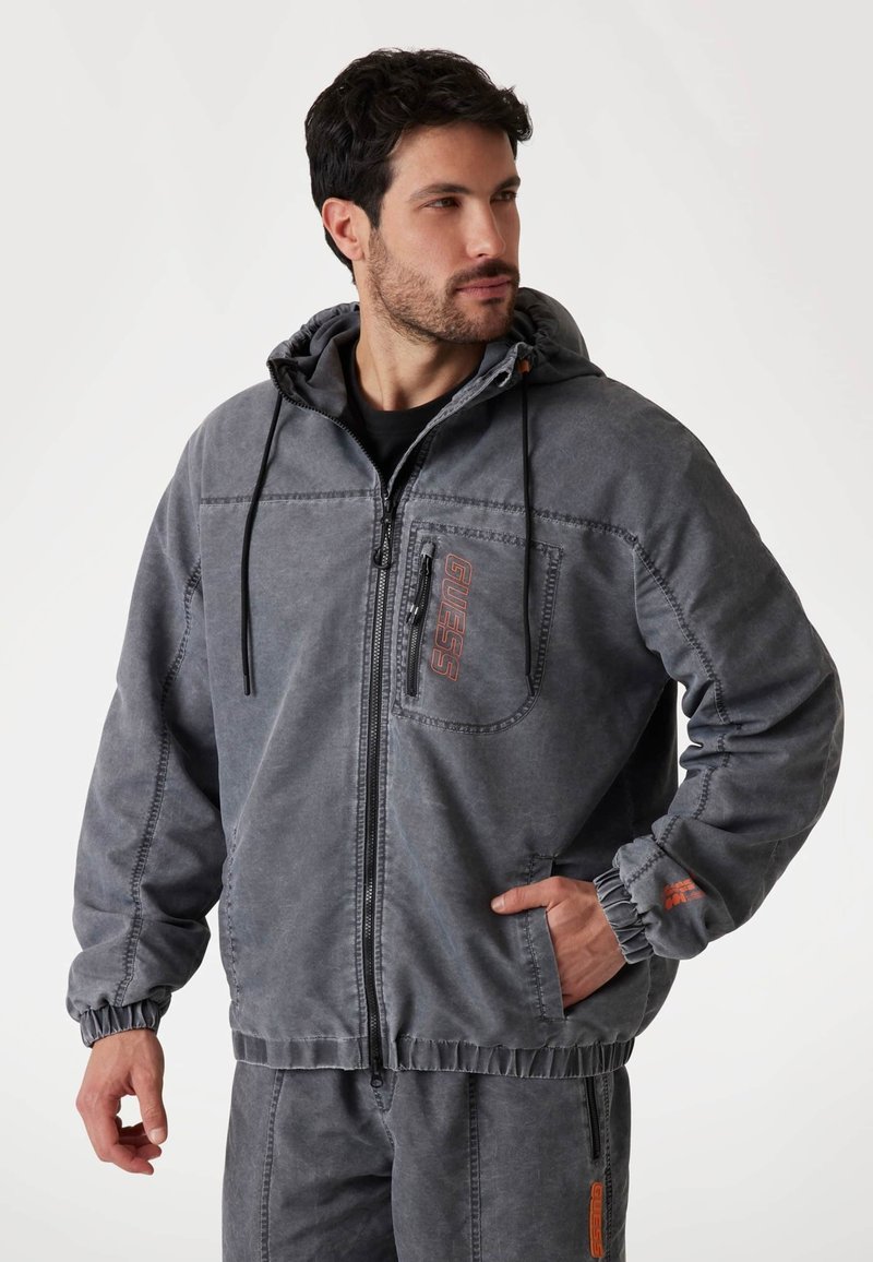 Chaqueta con capucha gris, con cierre de cremallera, puños elásticos y un bolsillo frontal. Presenta un detalle del logo en naranja y un acabado de tela texturizada.