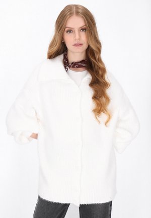 Weißer übergroßer Strickcardigan mit einer strukturierten Oberfläche, Knopfverschluss und umgeschlagenen Ärmeln, getragen über einem hellen Oberteil und kombiniert mit grauen Jeans.