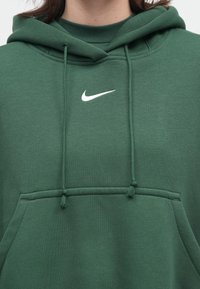 Nærbilde av en person som har på seg en mørkegrønn hettegenser med hvit Nike-logo, snorer og frontlomme synlig.