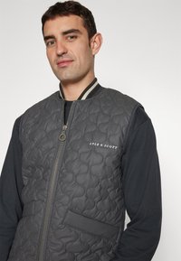 Quilted gray vest with a zip closure, ribbed collar, and a patch pocket. Features white text logo on the front. Worn over a dark shirt.