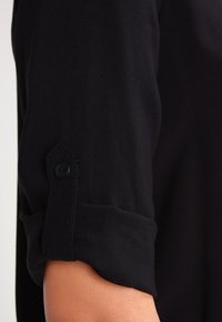 Sort langærmet skjorte med manchet rullet op og sikret med en sort knaplukning på en persons arm.