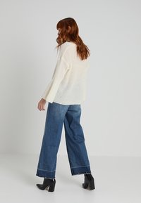 Pull en maille épaisse crème avec manches évasées, associé à un jean denim bleu à coupe ample avec ourlet effiloché, et des bottines noires.