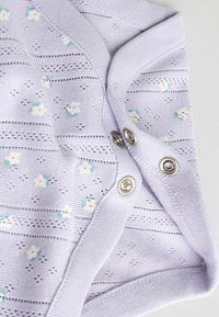 Lyse lilla strikket babybeklædning med små hvide og grønblå blomstermønstre og metaltrykknapper langs kanten.