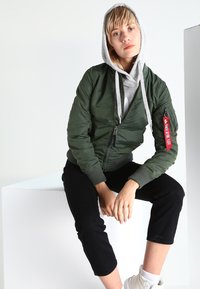 Zelená syntetická bomber bunda so zipsom vpredu, rebrovanými manžetami a červenou nášivkou. Kombinovaná so šedou mikinou a čiernymi skrátenými nohavicami. Biele tenisky.