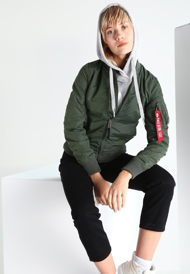 Zelená syntetická bomber bunda so zipsom vpredu, rebrovanými manžetami a červenou nášivkou. Kombinovaná so šedou mikinou a čiernymi skrátenými nohavicami. Biele tenisky.