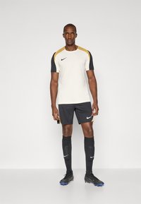 Nike sportssett med en kremfarget trøye med gull- og svart detaljer, kombinert med svarte shorts og sokker, samt svarte fotballsko.