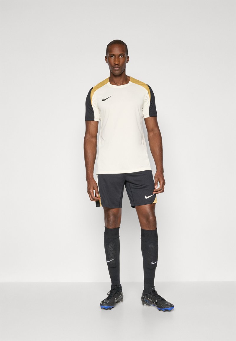 Vêtement de sport Nike comprenant un maillot crème avec des accents dorés et noirs, associé à un short noir, des chaussettes noires et des chaussures de football noires.