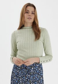 Helles grüne Rippstrickpullover mit einem Stehkragen und langen Ärmeln, verziert mit Knopfdetails an den Manschetten. Kombiniert mit einem rock mit floralen Mustern.