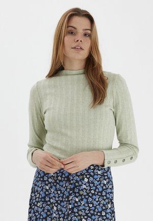 Maglione verde chiaro a coste con colletto alto e maniche lunghe, caratterizzato da dettagli in bottone sui polsini. Indossato con una gonna a fantasia floreale.