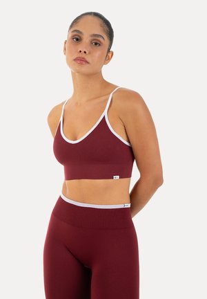 Mujer con top corto de canalé color granate y leggings de talle alto con ribete blanco, de pie con los brazos detrás de la espalda sobre un fondo liso.