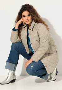 Chaqueta acolchada de color beige claro con dos bolsillos en el pecho, blusa de patrón azul, vaqueros azules arremangados en el tobillo y botas plateadas de tobillo.