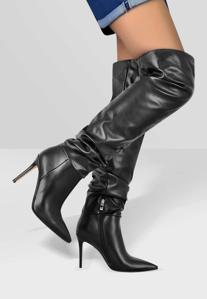 Jambes en jean bleu retroussé portant des bottes en cuir noires, à bout pointu, montantes jusqu'au genou avec des talons aiguilles hauts, sur fond gris.