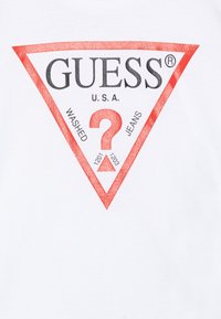 Weißes T-Shirt mit einem roten dreieckigen Logo, das den Text "GUESS" und "U.S.A." zeigt, ein großes Fragezeichen sowie die Worte "WASHED" und "JEANS."