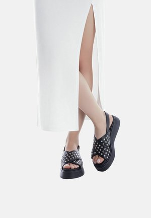 Sandalias negras con un diseño de tachuelas, punta abierta y suela gruesa. Llevadas con un vestido blanco que tiene una abertura en un lado.