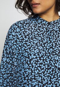 Blaue und schwarze Bluse mit Blumenmuster, Rundhalsausschnitt, leichter Stoff und einem Knopfdetail am Kragen.