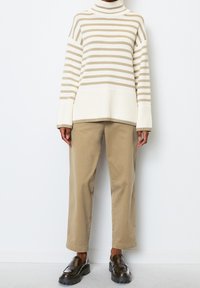 Rollkragenpullover mit cremefarbenen und beigen Streifen, breiten Ärmeln, kombiniert mit hellbraunen Hosen und dunkelbraunen Slippern.