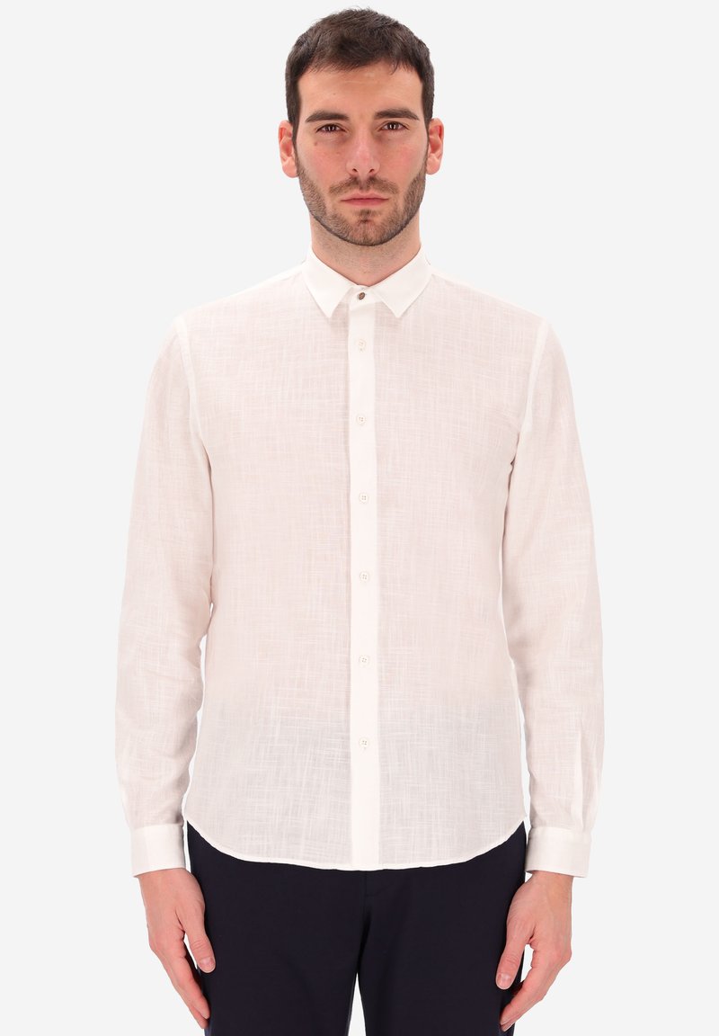 Uomo che indossa una camicia bianca a maniche lunghe con texture e pantaloni scuri, in piedi davanti a uno sfondo chiaro e uniforme.