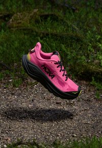 Lys rosa Hoka joggesko med svart såle og lisser som svever over en jordsti omgitt av grønt gress og løvverk.