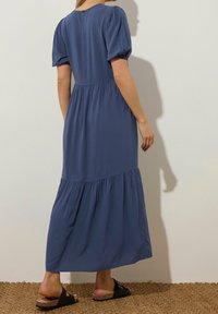 Robe maxi en tissu bleu avec des manches bouffantes, taille froncée et ourlet à volants. Le dos présente une fermeture à boutons. Le modèle porte des sandales à glissière noires.