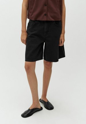 Person trägt lockere schwarze Denim-Shorts, dunkelbraunes ärmelloses Oberteil mit Knöpfen und schwarze Ballerinas, steht vor weißem Hintergrund.