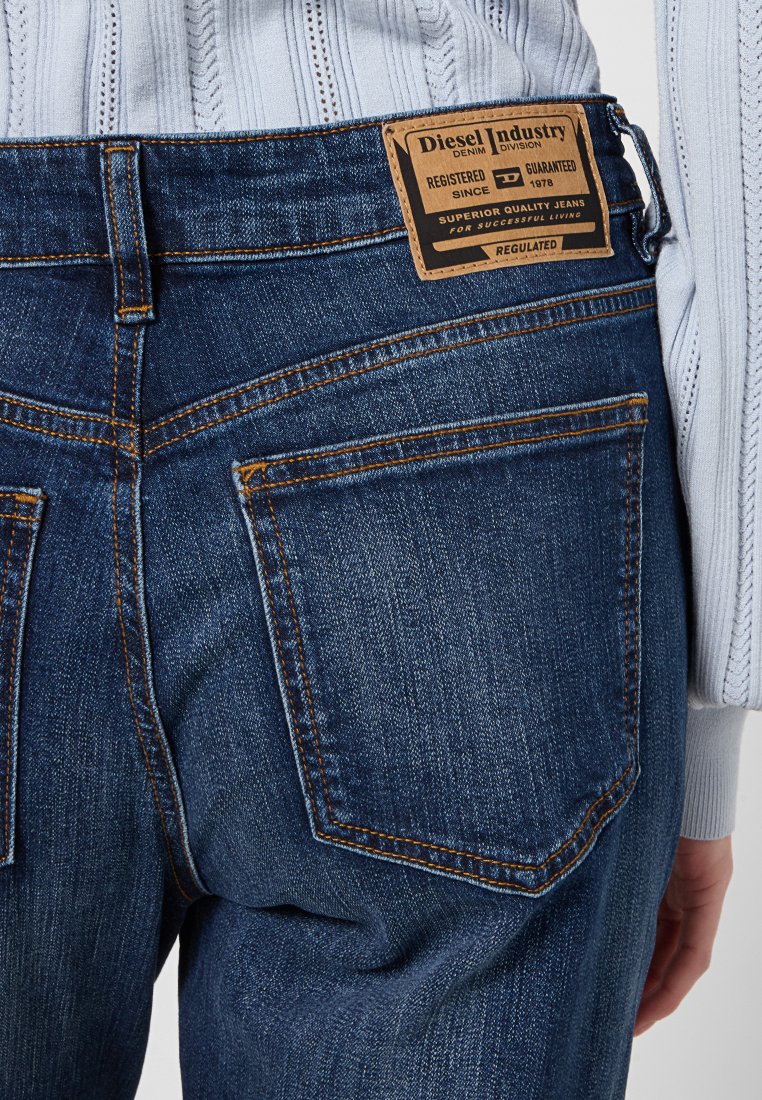 Jeans in denim blu con una toppa con il logo del marchio, due tasche posteriori, cuciture gialle e un cinturino testurizzato. Lavaggio scuro con una finitura liscia.