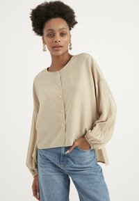 Helle beige Knopfleiste mit langen, voluminösen Ärmeln und elastischen Bündchen, aus einem glatten Stoff gefertigt. Kombiniert mit blauen Jeans.