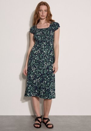 Femme portant une robe verte imprimée feuilles longueur genoux avec manches bouffantes et sandales noires à brides, debout contre un mur uni.