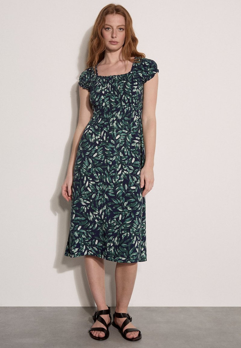 Femme portant une robe verte imprimée feuilles longueur genoux avec manches bouffantes et sandales noires à brides, debout contre un mur uni.