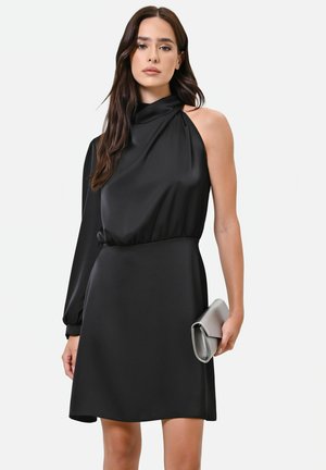 Femme aux longs cheveux foncés portant une robe noire asymétrique tenant une pochette argentée sur un fond clair uni.